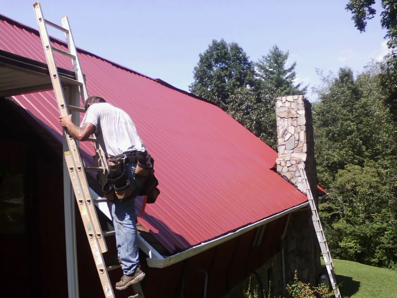 Roofer on a ladder beside a red metal roof and stone chimney — Soffit & Fascia Repair by Roofing Brothers in Kunkletown